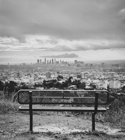 grayscale photo of bench near city buildings